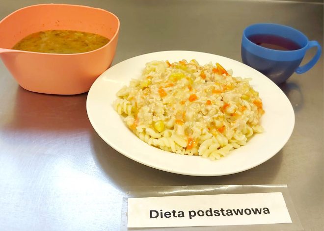 Porcja makaronu świderki z sosem warzywno-mięsnym oraz miseczka zupy jarzynowej. Do posiłku podano kubek z czerwonym kompotem. Pod talerzem kartka z podpisem „Dieta podstawowa”