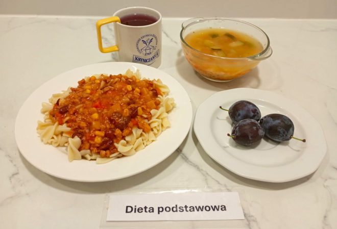 Na zdjęciu widać zestaw obiadowy w diecie podstawowej. Na głównym talerzu znajduje się makaron świderki z sosem bolońskim, w którym widać mięso mielone, kukurydzę, fasolę i warzywa. Obok podano przezroczystą miskę z zupą jarzynową oraz talerzyk z trzema śliwkami. Do posiłku dodano kubek z herbatą owocową