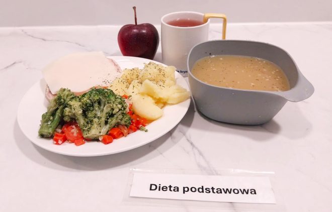 Obiad w diecie podstawowej: talerz z gotowanymi ziemniakami posypanymi koperkiem, warzywami – brokułem i marchewką oraz plasterkiem mięsa pod serem. Do tego jabłko, kubek z herbatą owocową i miska z zupą jarzynową na bulionie