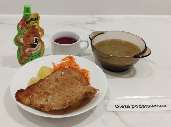 Talerz z obiadem: panierowany kotlet schabowy, ziemniaki i surówka z marchewki, obok kubek z kompotem, miseczka z zupą jarzynową oraz deser w saszetce. Podpis: „Dieta podstawowa”