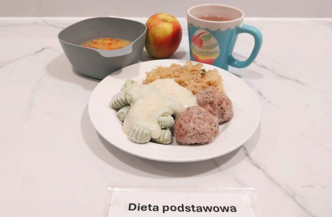 Na białym talerzu podane dwa pulpety z mięsa mielonego, obok zielone kopytka polane jasnym sosem oraz porcja duszonej kapusty. Z boku jabłko, szklanka herbaty w turkusowym kubku i miseczka z zupą jarzynową. Na pierwszym planie kartka z napisem „Dieta podstawowa”