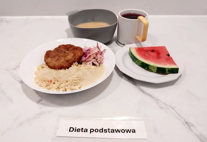 Na talerzu porcja ryżu polanego sosem, kotlet mielony panierowany i surówka z kapusty czerwonej. Obok miseczka z zupą kremem, kubek z herbatą oraz kawałek arbuza na osobnym talerzyku. Przed zestawem kartka z napisem „Dieta podstawowa”