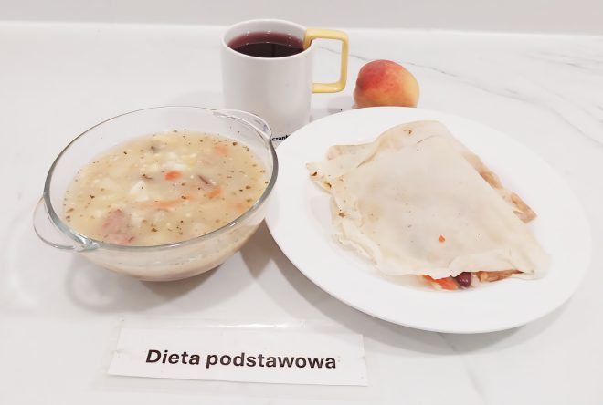 Zupa jarzynowa w szklanej miseczce, obok talerz z naleśnikiem nadziewanym warzywami. Do zestawu dołączony kubek z herbatą i brzoskwinia. Na stole karteczka z napisem „Dieta podstawowa”