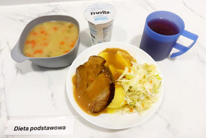 Talerz z pieczenią wieprzową w sosie, ziemniakami i surówką z białej kapusty. Obok miseczka z zupą jarzynową, kubek z herbatą i jogurt naturalny. Na stole leży kartka z napisem „Dieta podstawowa”