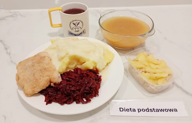 Taca z obiadem w diecie podstawowej: na talerzu panierowany filet rybny, puree ziemniaczane z sosem, buraczki. Obok miseczka zupy, kompot w kubku oraz deser – kasza jęczmienna z gotowanymi jabłkami