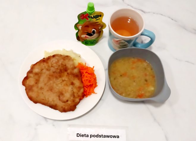 Obiad w ramach diety podstawowej. Na talerzu znajduje się kotlet schabowy w panierce, podany z puree ziemniaczanym oraz surówką z marchwi. W miseczce podano zupę jarzynową na wywarze warzywnym. Do zestawu dołączono kubek herbaty oraz mus owocowy w tubce