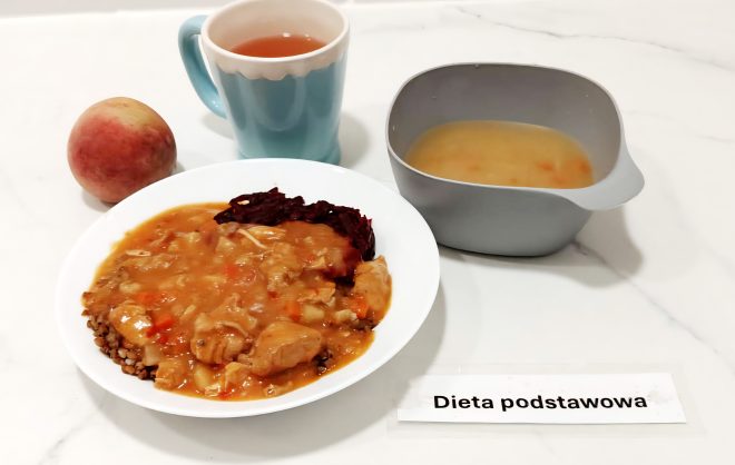 Na białym talerzu porcja kaszy gryczanej z gulaszem drobiowym w sosie warzywnym i surówką z buraków. Obok miseczka z zupą jarzynową, kubek z herbatą oraz brzoskwinia. Na stole karteczka z napisem „Dieta podstawowa”