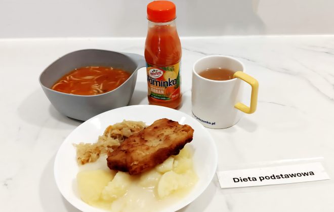 Na białym talerzu porcja puree ziemniaczanego z kapustą zasmażaną i smażonym filetem rybnym. Obok miseczka z zupą pomidorową z makaronem, kubek z herbatą oraz butelka napoju owocowego „Vitamina Hortex”. Na stole karteczka z napisem „Dieta podstawowa”