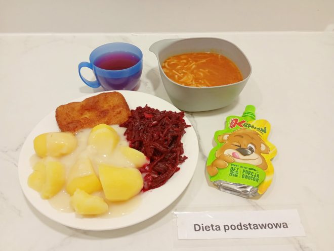Na białym talerzu znajdują się ziemniaki polane sosem, panierowany filet rybny oraz buraczki na ciepło. Obok stoi miska z zupą pomidorową z makaronem, niebieski kubek z herbatą oraz mus owocowy w saszetce dla dzieci