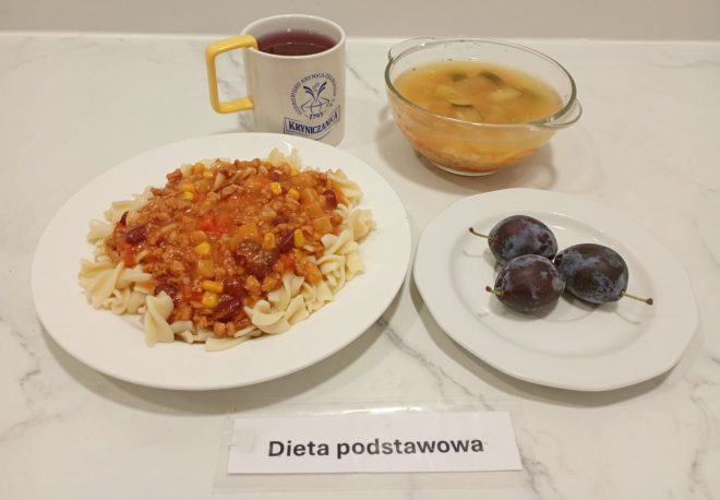 Na zdjęciu zestaw obiadowy w diecie podstawowej. Na głównym talerzu znajduje się porcja makaronu świderki z sosem mięsno-warzywnym, zawierającym m.in. kukurydzę, fasolę i pomidory. Obok ustawiono przezroczystą miskę z zupą jarzynową oraz talerzyk z trzema śliwkami. Do posiłku podano kubek z czerwoną herbatą owocową