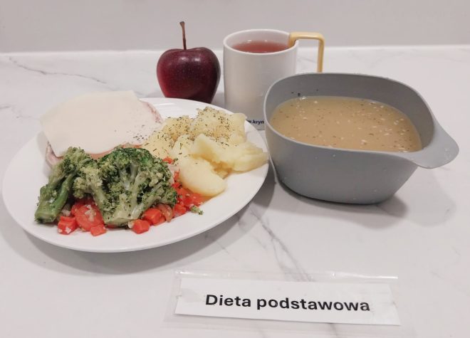 Obiad w diecie podstawowej: talerz z gotowanymi ziemniakami, brokułem i marchewką, plasterkiem mięsa z serem, a także jabłko. Obok znajduje się miseczka z zupą jarzynową oraz kubek z herbatą