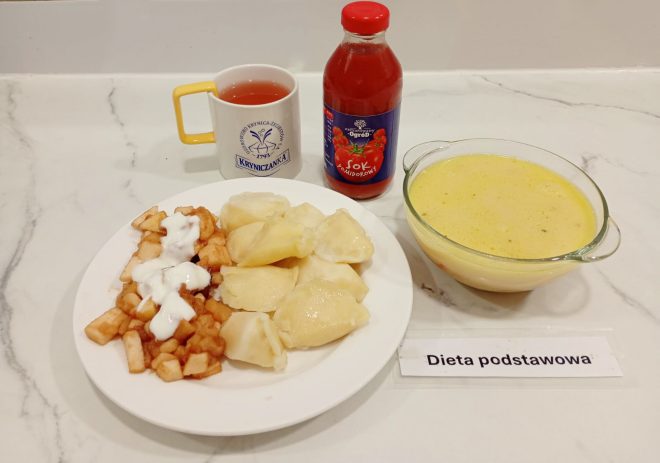 Zdjęcie obiadu w diecie podstawowej: talerz z pierogami podanymi z duszonymi jabłkami polanymi jogurtem, obok kubek z herbatą owocową i butelka soku pomidorowego. Po prawej stronie miska z żółtą zupą kremem