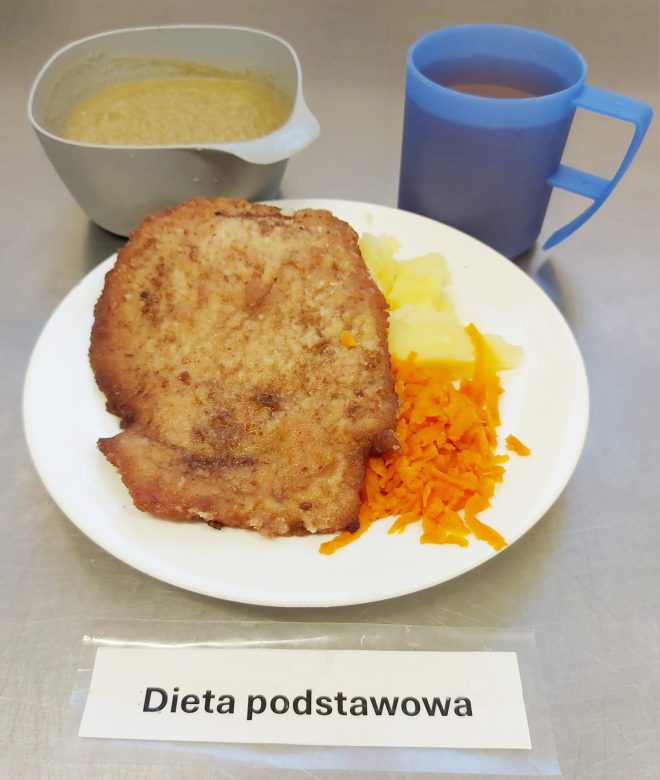 Na zdjęciu widoczny jest talerz z obiadem w ramach „diety podstawowej”. Na talerzu znajduje się panierowany kotlet schabowy, porcja gotowanych ziemniaków oraz surówka z marchwi. W tle ustawiono miseczkę z zupą krem oraz niebieski kubek z herbatą. Przed talerzem leży karteczka z napisem „Dieta podstawowa”