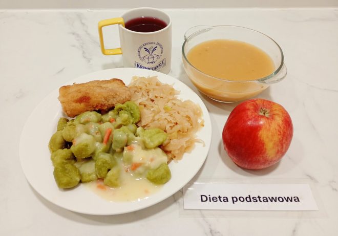 Na talerzu znajduje się porcja zielonych klusek polanych jasnym sosem z warzywami, smażony filet rybny oraz kapusta kiszona. Obok ułożone są: jabłko, miseczka z zupą oraz kubek z czerwonym napojem. Przy posiłku leży karteczka z napisem „Dieta podstawowa”