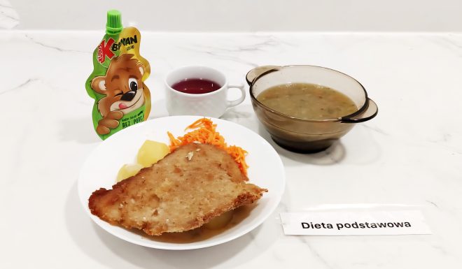 Talerz z panierowanym kotletem, ziemniakami i surówką z marchewki. Obok znajduje się miseczka z zupą jarzynową, kubek z herbatą oraz mus owocowy w saszetce dla dzieci. Na stole widoczna jest karteczka z napisem „Dieta podstawowa”