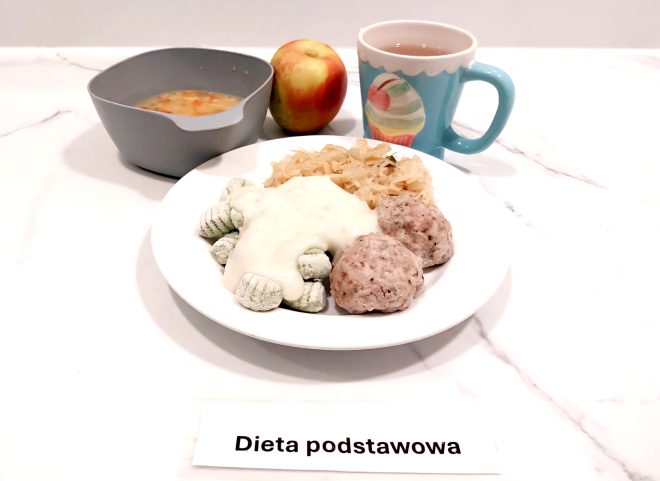 Na tacy znajduje się obiad składający się z talerza z zielonymi kluskami gnocchi polanymi białym sosem, dwóch pulpecików z mięsa mielonego i porcji duszonej kapusty. W miseczce podano zupę jarzynową, a jako dodatek owoc – jabłko. Do picia przygotowano kubek herbaty. Na tacy widoczna jest etykieta „Dieta podstawowa”