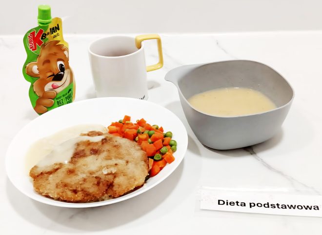 Na białym stole ustawiono zestaw obiadowy z napisem „Dieta podstawowa”. Posiłek składa się z zupy krem w szarej miseczce, panierowanego kotleta z sosem, gotowanej marchewki z groszkiem, a do tego podano napój w kubku oraz deser w formie owocowego musiku w tubce z wizerunkiem misia