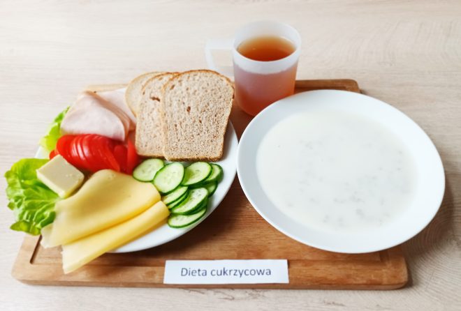 Na tacy ustawiono zestaw śniadaniowy. Po lewej stronie znajduje się talerz z trzema kromkami pieczywa pełnoziarnistego, plastrem szynki drobiowej, dwiema porcjami żółtego sera, kostką białego sera, liściem sałaty, plastrami pomidora i świeżego ogórka. Obok, w białej misce, podano zupę mleczną z drobnymi dodatkami. Całość uzupełnia kubek z herbatą bez cukru. Na tacy widoczna jest etykieta „Dieta cukrzycowa”
