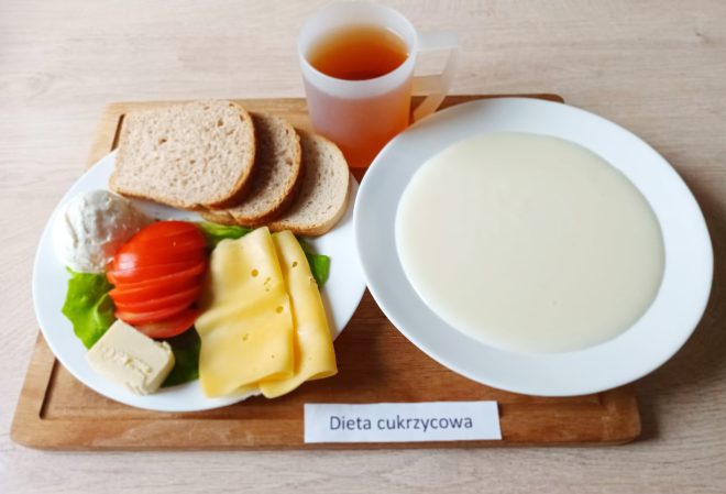 Śniadanie w diecie cukrzycowej: kromki pieczywa razowego, plasterki sera żółtego, pomidor, liść sałaty, twaróg i margaryna. Obok talerz z kaszką mleczną i kubek z herbatą owocową