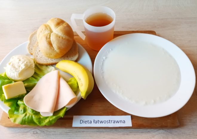 Na tacy ułożono śniadanie przygotowane dla osób na diecie łatwostrawnej. Na talerzu znajduje się bułka kajzerka, kromka chleba pszennego i kromka chleba graham, plaster sera żółtego, plaster wędliny drobiowej, liść sałaty, kulka twarogu oraz banan. Obok ustawiono drugi talerz z talerzem białej, kremowej zupy mlecznej. Do posiłku podano także kubek z jasnobrązowym napojem, prawdopodobnie herbatą