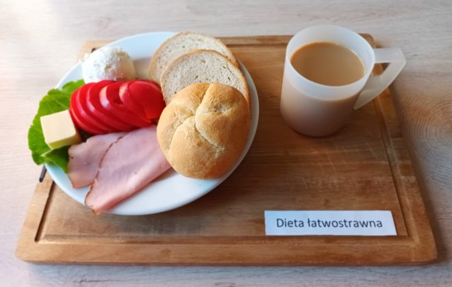 Zestaw śniadaniowy w diecie łatwostrawnej: bułka i kromki chleba, plasterki szynki, pomidor, kostka sera żółtego, kulka twarogu, liść sałaty oraz kubek kawy z mlekiem