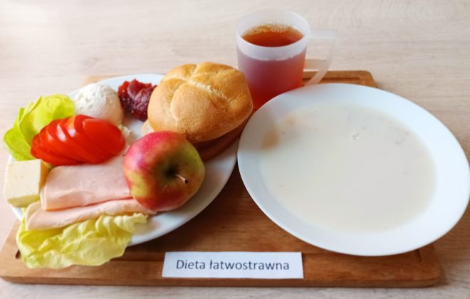 Na drewnianej tacy znajduje się posiłek oznaczony jako „Dieta łatwostrawna”. Na białym talerzu widać plasterki pomidora, liść sałaty, ser biały, plasterki szynki, kostkę sera żółtego, porcję dżemu, bułkę pszenną i jabłko. Obok stoi kubek z herbatą oraz miska z jasną, kremową zupą
