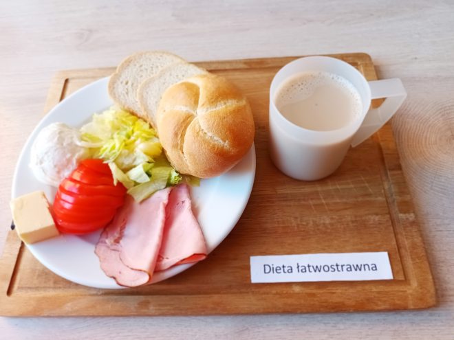 Śniadanie w diecie łatwostrawnej: na talerzu pieczywo pszenne – bułka i kromki chleba, obok plasterek sera żółtego, plasterki szynki drobiowej, pomidor, sałata i twarożek. Do posiłku podano kubek z kawą z mlekiem lub kawą zbożową