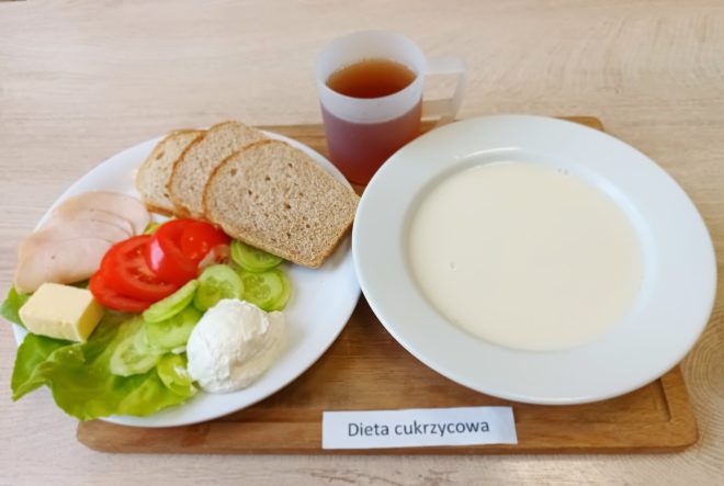 Na zdjęciu przedstawiono zestaw śniadaniowy z podpisem „Dieta cukrzycowa”. Na tacy znajduje się talerz z trzema kromkami pieczywa pełnoziarnistego, plasterkami pomidora i ogórka, sałatą, kawałkiem masła, porcją twarożku oraz plastrami drobiowej wędliny. Obok stoi miska z mlekiem oraz kubek z herbatą