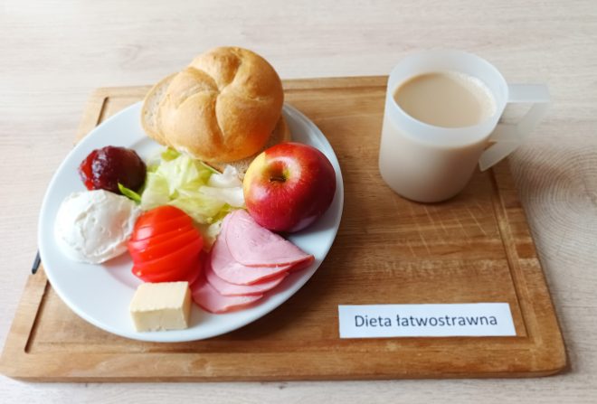 Na białym talerzu podano bułkę kajzerkę, kilka plasterków szynki, pomidor, liście sałaty, serek twarogowy, kostkę żółtego sera, porcję dżemu oraz jabłko. Towarzyszy im kubek kawy z mlekiem. Całość ustawiona jest na drewnianej tacy z karteczką „Dieta łatwostrawna”