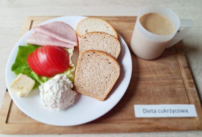 Na drewnianej tacy umieszczono talerz śniadaniowy z napisem „Dieta cukrzycowa”. Na talerzu znajdują się trzy kromki chleba pszenno-żytniego, plasterki wędliny, pomidor z listkiem sałaty, porcja twarogu, kawałek masła, a obok w plastikowym kubku podano kawę z mlekiem