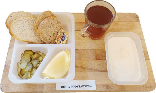 Na drewnianej tacy ułożono śniadanie w diecie podstawowej: trzy kromki pieczywa, porcję masła w złotym opakowaniu, plaster sera żółtego, ogórki konserwowe, kubek z ciemną herbatą oraz plastikowy pojemniczek z jogurtem naturalnym