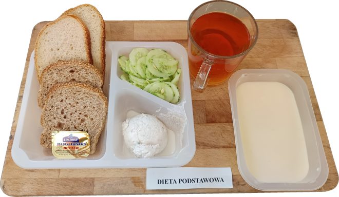 Zestaw śniadaniowy w diecie podstawowej: kilka kromek chleba jasnego i pełnoziarnistego, świeży ogórek w plasterkach, kulka twarogu, porcja masła, jogurt naturalny w miseczce oraz herbata w szklance