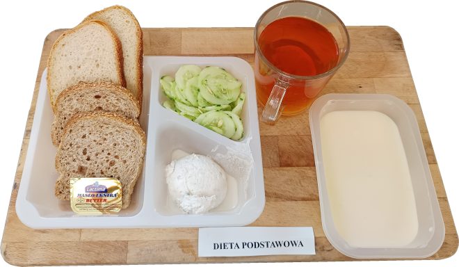 Śniadanie w ramach diety podstawowej serwowane w plastikowej zastawie. Na talerzu znajdują się trzy kromki pieczywa pszenno-żytniego, kostka masła, kulka twarożku oraz plasterki świeżego ogórka. Obok ustawiono pojemniczek z jogurtem naturalnym i szklankę herbaty