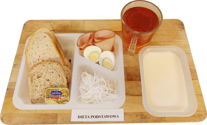 Na drewnianej tacy ustawiono zestaw śniadaniowy opisany jako „Dieta podstawowa”. W plastikowym talerzu podzielonym na przegródki znajdują się kromki chleba pszennego i żytniego wraz z porcją masła w kostce, plasterki szynki drobiowej, połówki gotowanego jajka oraz surówka z białej kapusty. Obok podano porcję zupy mlecznej w prostokątnym pojemniku oraz szklankę herbaty