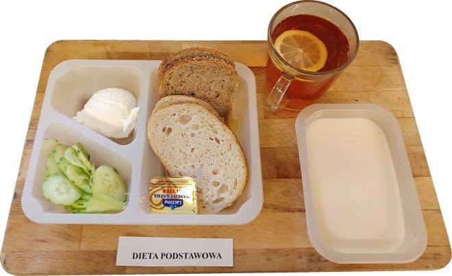 Posiłek w pojemniku porcjowym: twaróg, świeży ogórek i pieczywo mieszane z dodatkiem masła. Do zestawu szklanka herbaty z cytryną oraz miseczka jogurtu lub mleka. Na tacy kartka z podpisem „Dieta podstawowa”