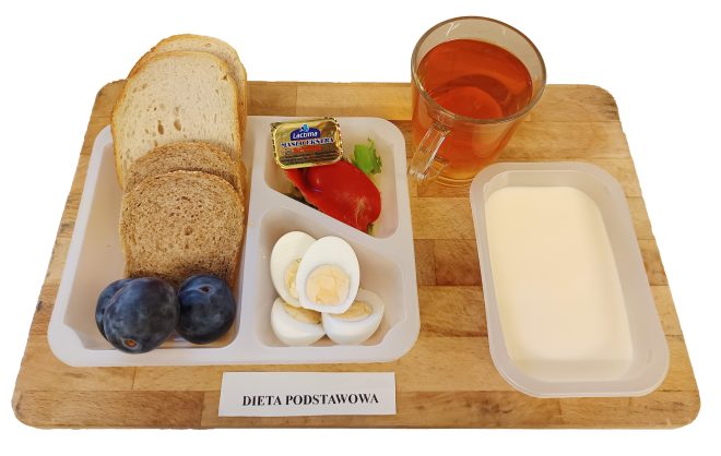 Na tacy podano pieczywo jasne i ciemne, gotowane jajko pokrojone w plastry, dwie śliwki, plaster papryki z listkiem sałaty oraz porcję masła. Obok stoi szklanka herbaty oraz pojemniczek z jasnym deserem mlecznym