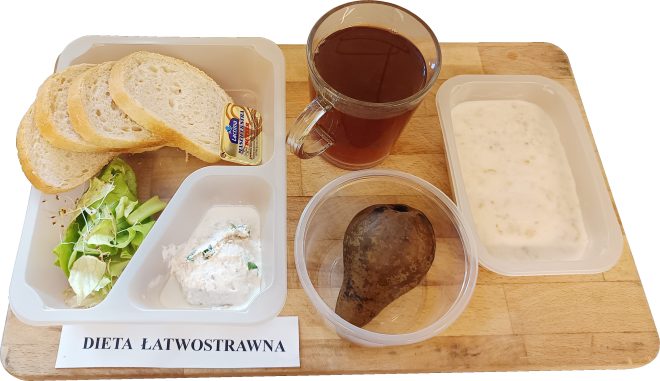 Taca ze śniadaniem w diecie łatwostrawnej: w pojemniku kilka kromek jasnego pieczywa i porcjonowane masło, obok sałata z kiełkami oraz pasta twarogowa. Na tacy dodatkowo znajduje się gotowana gruszka w pojemniku, kubek z herbatą i miseczka z mleczną owsianką