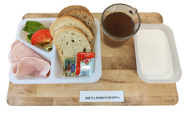 Śniadanie w diecie podstawowej: zestaw na plastikowej tacce – plasterki szynki drobiowej, liść sałaty, pomidor, porcje masła i dżemu truskawkowego oraz kilka kromek chleba pszenno-żytniego. Do tego szklanka kawy zbożowej i miseczka z jogurtem naturalnym
