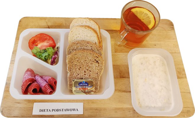 Na tacy przygotowano zestaw śniadaniowy z etykietą „Dieta podstawowa”. Składa się z kilku kromek pieczywa jasnego i razowego, porcjowanego masła, plasterków salami, kawałka pomidora i liści sałaty. Obok stoi kubek herbaty z cytryną oraz miseczka z owsianką na mleku.