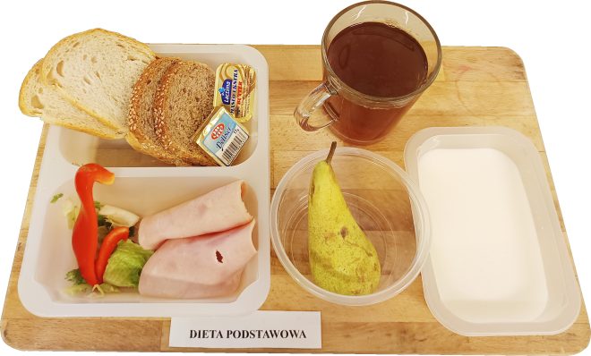 Na tacy ułożono posiłek z napisem „Dieta podstawowa”. W zestawie są kromki pieczywa jasnego i razowego, dwa rodzaje masła w porcjach, plasterki szynki, kawałki sałaty i papryki, gruszka, szklanka kawy zbożowej oraz jogurt naturalny w plastikowym pojemniku.