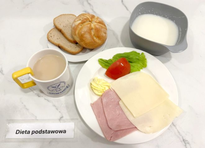 Na talerzu dwa plasterki szynki, dwa plasterki sera żółtego, liść sałaty i kawałek pomidora, a także porcja masła. Obok miseczka z jogurtem naturalnym, pieczywo (kajzerka i kromki chleba) oraz kubek kawy z mlekiem. Na pierwszym planie kartka z napisem „Dieta podstawowa”