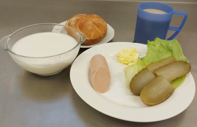 Śniadanie: bułka kajzerka, porcja pasztetu drobiowego, plasterki ogórka kiszonego na liściu sałaty, masło, miseczka z mlekiem i kubek kawy z mlekiem
