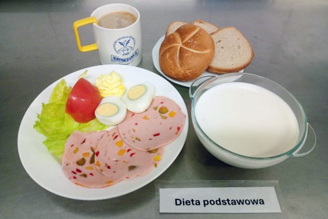Śniadanie w diecie podstawowej: talerz z liściem sałaty, plasterkiem pomidora, porcją margaryny, połową jajka na twardo oraz kilkoma plastrami mortadeli z papryką. Do tego bułka i kromki chleba, miseczka zupy mlecznej i kubek kawy zbożowej