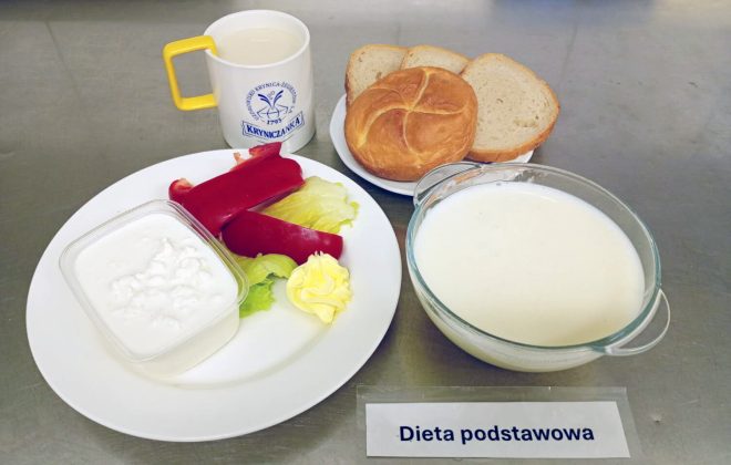 Śniadanie w diecie podstawowej: pieczywo jasne i bułka, serek wiejski w miseczce, papryka czerwona i liście sałaty, porcja masła. Obok miseczka zupy mlecznej oraz kubek kawy zbożowej z mlekiem