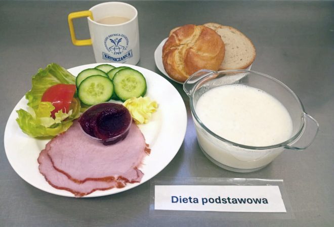 Zestaw śniadaniowy w diecie podstawowej: trzy kromki chleba pszennego, plasterki szynki, sałatka z pomidora, ogórka i sałaty, porcja masła, kawa z mlekiem podana w kubku „Kryniczanka”