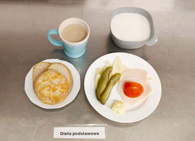 Na tacy podano śniadanie dla osób na diecie podstawowej. Na talerzu znajdują się kromki chleba pszennego i bułka kajzerka. Obok, na drugim talerzu, podano plaster sera żółtego, plaster wędliny drobiowej, kawałek pomidora, dwa ogórki kiszone, listki sałaty oraz porcję masła. Do zestawu dołączono miseczkę z mlekiem oraz kubek kawy z mlekiem