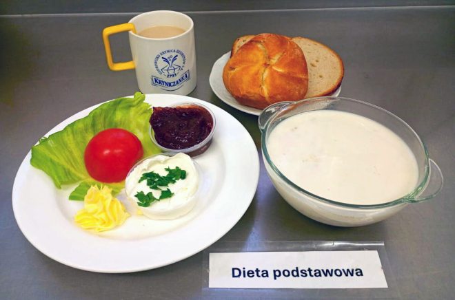 Na tacy znajduje się zestaw śniadaniowy opisany jako „Dieta podstawowa”. Na białym talerzu ułożono liść sałaty z całym pomidorem, porcję twarożku posypanego natką pietruszki, masło oraz mały kubeczek z dżemem. Obok podano bułkę pszenną i kromki chleba jasnego, a także miseczkę z zupą mleczną. Do zestawu dołączono kubek kawy z mlekiem w filiżance z logo „Kryniczanka”