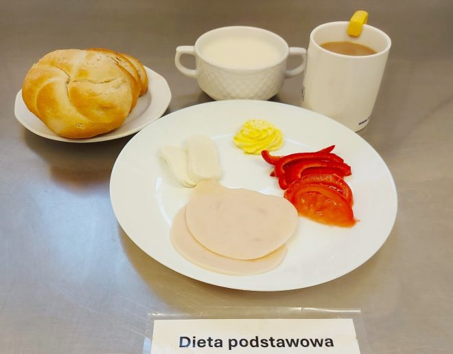 Talerz z dwoma plasterkami wędliny drobiowej, kawałkiem sera białego, plastrem pomidora i paskami papryki czerwonej, porcją masła oraz pieczywem: bułką i kromkami chleba. Do tego kubek kawy z mlekiem i miseczka mleka. Pod talerzem kartka z napisem „Dieta podstawowa”