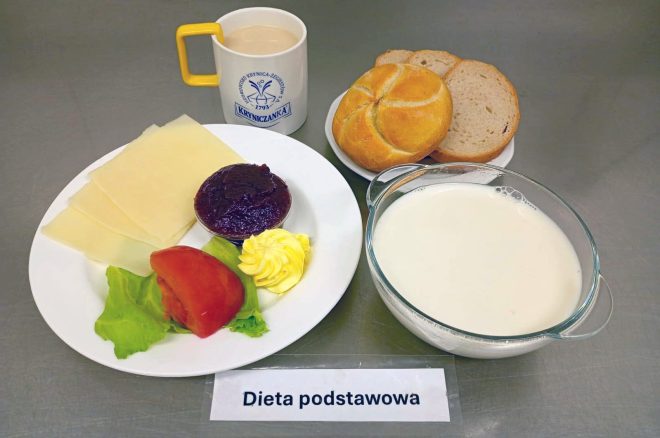 Na zdjęciu przedstawiono śniadanie w diecie podstawowej. Na talerzu leży kilka plastrów sera żółtego, liść sałaty, kawałek pomidora, porcja masła oraz porcja konfitury. Obok znajduje się talerzyk z bułką kajzerką i kromkami chleba. Do zestawu dołączono kubek z kawą z mlekiem oraz miseczkę z mlekiem