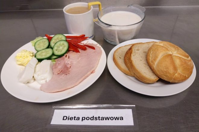 Śniadanie w diecie podstawowej: talerz z plasterkami szynki drobiowej, świeżym ogórkiem i papryką, porcją masła i białego sera. Obok leżą kromki chleba pszennego i bułka. Do posiłku podano kubek z kawą z mlekiem oraz miseczkę jogurtu naturalnego